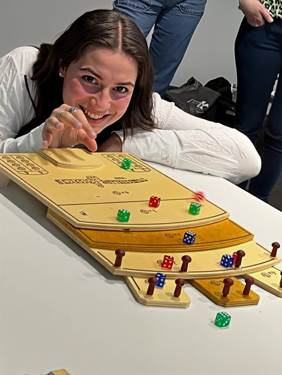 Teilnehmerin schnippt einen Würfel über ein hölzernes Spielbrett, Mitspielende warten daneben